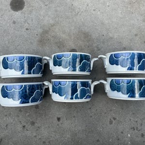 Op de afbeelding: Zes witte keramische kommen met blauwe wolkenpatronen en kleine handvatten. De kommen zijn gerangschikt in twee rijen van drie, wat een samenhangend ontwerp laat zien. Ideaal voor het serveren van soepen of kleine gerechten.