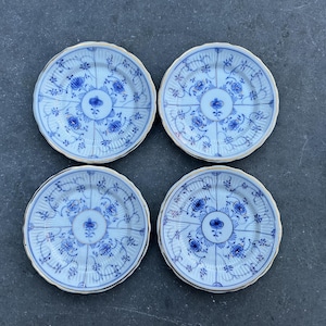 Puede incluir: Cuatro platos pequeños redondos de porcelana azul y blanca con un diseño floral. Los platos tienen un borde festoneado y un borde dorado. Los platos están dispuestos en forma cuadrada sobre una superficie oscura. Hay platos adicionales apilados a la derecha.
