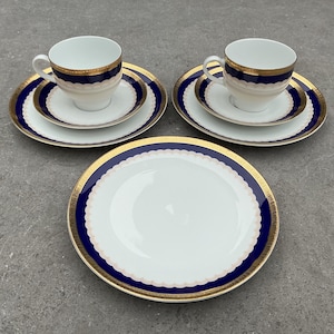 Könnte beinhalten: Ein weißes Porzellan-Teeservice mit gold- und marineblauen Verzierungen. Das Set enthält zwei Tassen und Untertassen, zwei kleinere Teller und einen größeren Teller. Die Teller haben einen gewellten Rand.