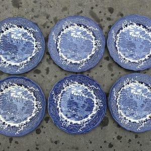 May include: Six blue and white decorative plates with a scalloped edge. Each plate features a detailed landscape scene with a castle, trees, and a bridge. The plates are arranged on a gray surface.