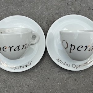 Può includere: Due tazze da espresso e piattini in ceramica bianca. Le tazze hanno la scritta "Operandi" stampata in argento, con "Modus Operandi" stampato sui piattini. Le tazze hanno un manico.