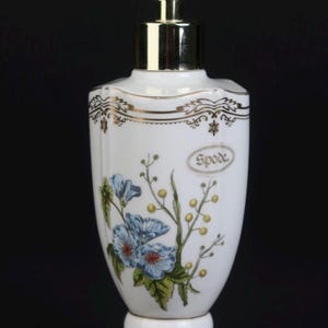 Op de afbeelding: Witte keramische zeepdispenser met een gouden pomp en voet. De dispenser heeft een bloemenontwerp met blauwe bloemen en het woord "Spode". Decoratieve gouden randen accentueren de bovenkant.