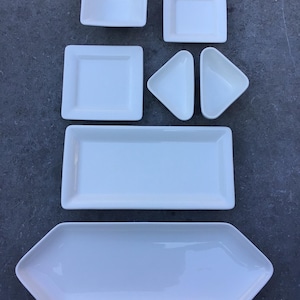 May include: A set of white ceramic serving dishes in various shapes and sizes, including a rectangular platter, a hexagonal platter, a square dish, and two triangular dishes.