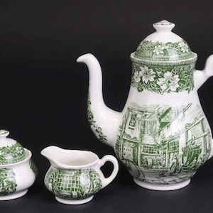 May include: A white ceramic tea set with a green floral and town scene design. The set includes a teapot, a sugar bowl with a lid, and a creamer. The teapot has a curved spout and handle.