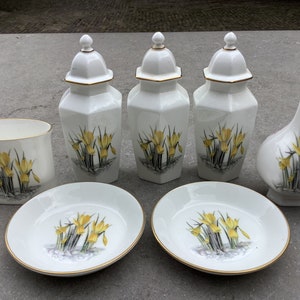 May include: A set of white porcelain kitchenware featuring a floral design with yellow daffodils. The set includes three jars, a vase, a small bowl, and a larger bowl.