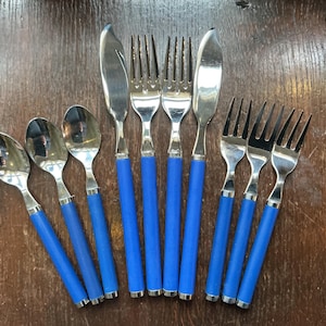 Puede incluir: Un juego de cubiertos de acero inoxidable con mangos azul brillante. El conjunto incluye cucharas, tenedores y cuchillos, dispuestos sobre una superficie de madera. Los cubiertos tienen un diseño moderno, con un acabado pulido.