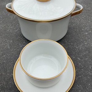 May include: White ceramic cookware set with gold trim. The set includes a lidded pot with handles and a small bowl with a saucer. The gold trim accents the edges of the pot, lid, bowl, and saucer. The set is designed for cooking and serving.