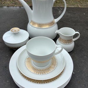 May include: A white porcelain tea set with gold accents. The set includes a teapot, creamer, sugar bowl with lid, and a cup and saucer. The set features a repeating pattern of stylised leaves and ovals.