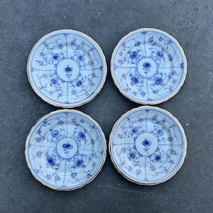 Puede incluir: Cuatro platos pequeños y redondos de porcelana azul y blanca con un diseño floral. Cada plato tiene un borde festoneado y un borde dorado. Los platos están dispuestos en forma cuadrada sobre una superficie gris oscuro.