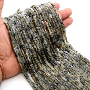 May include: A collection of rectangular beads in grey and black hues. The beads are strung together, forming a textured pattern. The beads are held in a hand against a white backdrop. The beads appear to be made of stone.