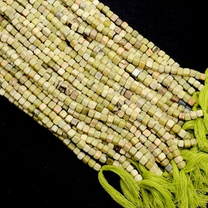 May include: Close-up of strands of small, square beads in shades of green, beige, and brown. The beads are strung together, creating a textured surface. A bundle of green thread is visible. The beads appear to be made of stone or a similar material.