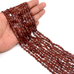 May include: Strands of triangular, dark red gemstone beads. The beads are strung together, showcasing their geometric shape and rich color. The beads are held in a hand against a white background.