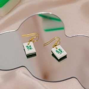 May include: A pair of gold earrings with green and white mahjong tiles. The tiles have Chinese characters on them.