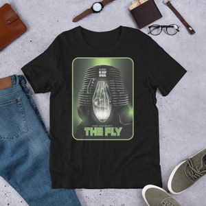 Puede incluir: Camiseta negra con un diseño de cartel de película de "The Fly". El cartel tiene una combinación de colores verde y gris con el texto "BE VERY AFRAID" y "DAVID CRONENBERG'S THE FLY".