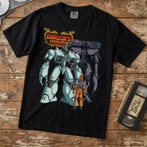 May include: Black t-shirt featuring a graphic of a white and gray robot with a person in an orange suit. The text "Dragon's Heaven" is above the robot in gold lettering. A VHS tape labeled "Retro Replay" is on the right.