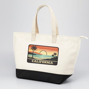 Peut inclure: Un sac fourre-tout en toile couleur crème avec deux anses et une base noire. L'avant présente un écusson rectangulaire illustrant un coucher de soleil sur une plage avec des palmiers et le mot CALIFORNIA en lettres majuscules claires.