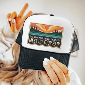 Può includere: Un cappellino trucker bianco e nero con una toppa rettangolare raffigurante un paesaggio in stile retrò con sole, alberi e acqua. La toppa include il testo: The best things in life MESS UP YOUR HAIR.