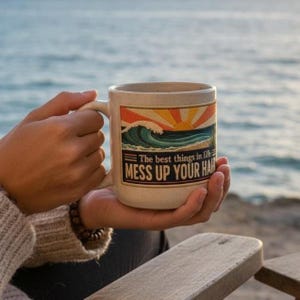 May include: A speckled off-white ceramic mug featuring a rectangular graphic of a stylized ocean wave and a rising sun in shades of orange, yellow, and blue. The text reads, The best things in life MESS UP YOUR HAIR in bold black lettering.