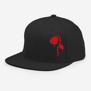 May include: A black baseball cap featuring an embroidered red rose design on the front panel. The rose graphic depicts a single flower with a stem and leaves, with red liquid-like drips extending downward from the petals and the leaf.