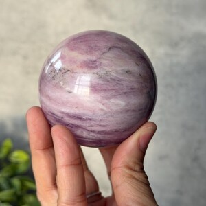May include: A polished spherical stone featuring a smooth surface with swirling patterns of light pink, lavender, and white, accented by subtle dark grey mineral inclusions throughout the rounded form of the natural gemstone specimen.