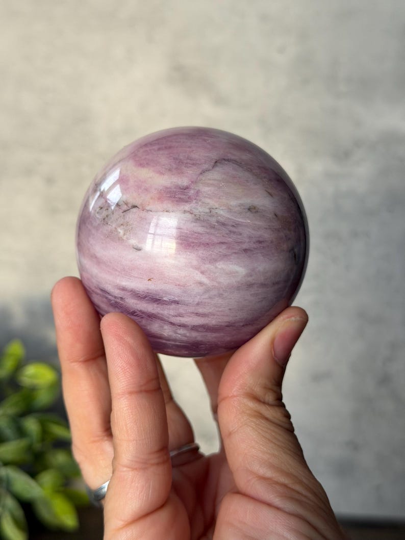 May include: A polished spherical stone featuring a smooth surface with swirling patterns of light pink, lavender, and white, accented by subtle dark grey mineral inclusions throughout the rounded form of the natural gemstone specimen.