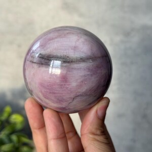May include: A polished spherical stone featuring a smooth surface with swirling patterns of light and dark pink hues, accented by thin, horizontal charcoal-coloured mineral inclusions that wrap around the centre of the orb.