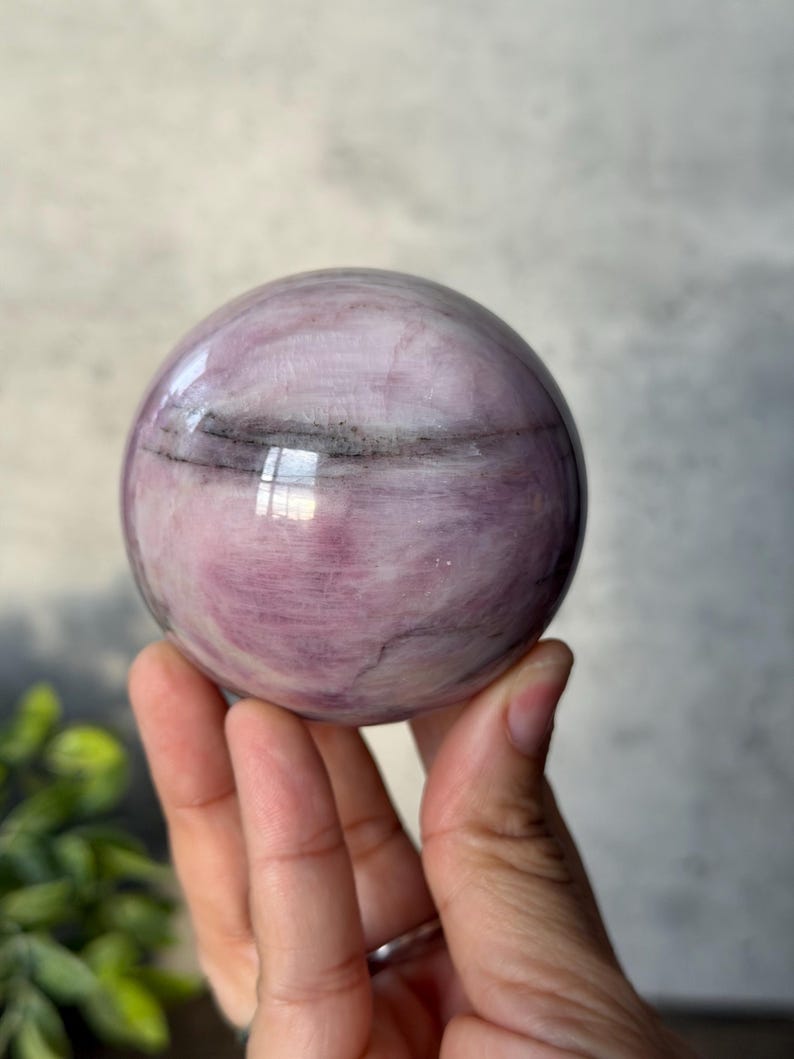 May include: A polished spherical stone featuring a smooth surface with swirling patterns of light and dark pink hues, accented by thin, horizontal charcoal-coloured mineral inclusions that wrap around the centre of the orb.