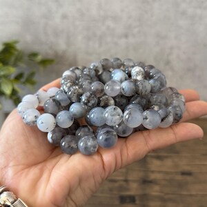 May include: A stack of gray and white gemstone bracelets with black flecks. The bracelets are made of round beads and are stacked on top of each other.