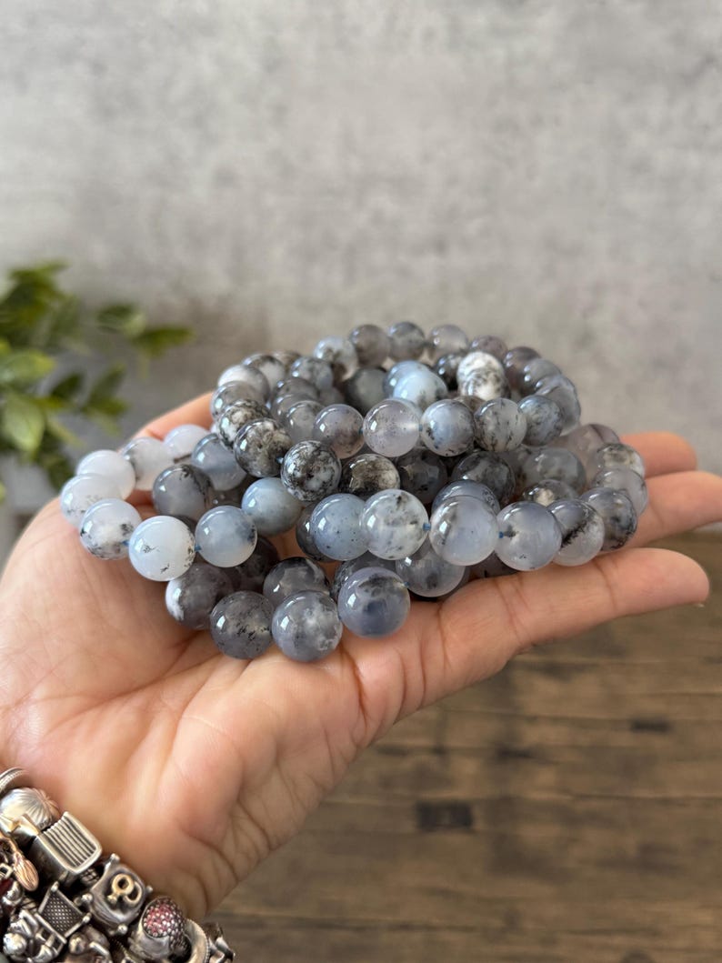 May include: A stack of gray and white gemstone bracelets with black flecks. The bracelets are made of round beads and are stacked on top of each other.
