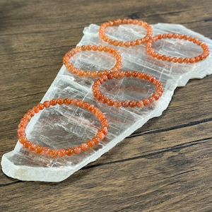 May include: Four orange sunstone bracelets on a clear crystal surface. The bracelets are made of small, round beads.