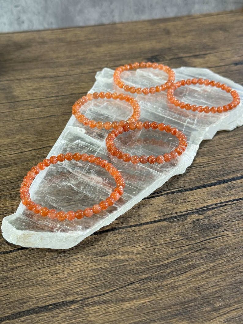 May include: Four orange sunstone bracelets on a clear crystal surface. The bracelets are made of small, round beads.