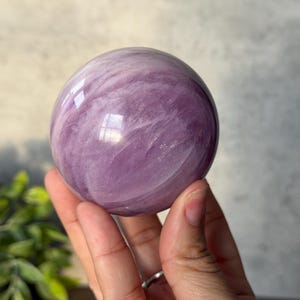 May include: A polished, spherical lepidolite crystal displaying a smooth, glossy surface with swirling patterns of light and deep lavender purple hues. The stone features natural internal inclusions and reflective highlights across its rounded form.