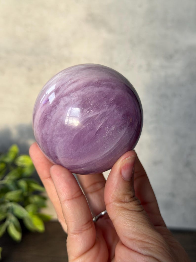 May include: A polished, spherical lepidolite crystal displaying a smooth, glossy surface with swirling patterns of light and deep lavender purple hues. The stone features natural internal inclusions and reflective highlights across its rounded form.