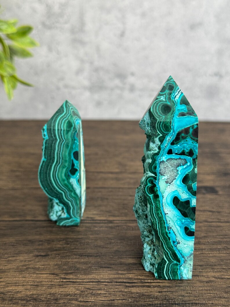 Puede incluir: Dos torres de cristal de azurita malaquita verde y azul sobre una superficie de madera. Las torres son puntiagudas en la parte superior y tienen una textura natural y rugosa.