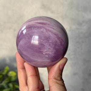 May include: A polished, spherical gemstone with a smooth, glossy surface. The stone features a translucent, light purple colour with swirling, cloud-like white inclusions throughout its structure, creating a marbled appearance with light reflections.