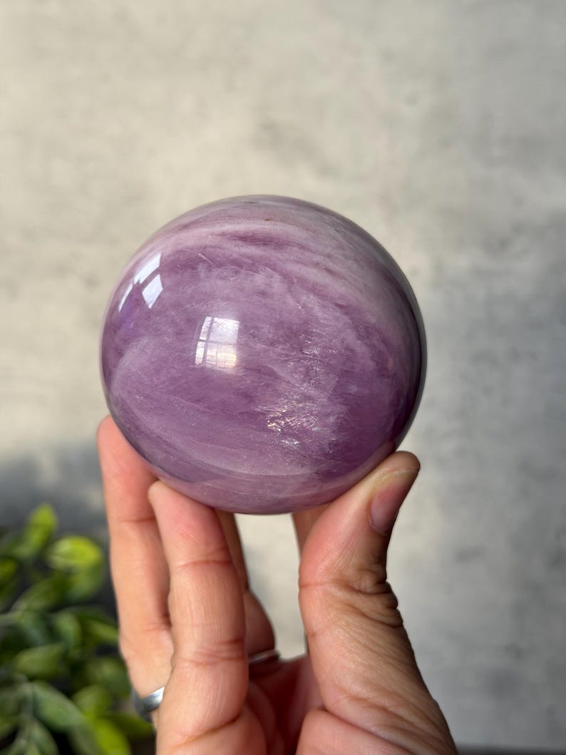 May include: A polished, spherical gemstone with a smooth, glossy surface. The stone features a translucent, light purple colour with swirling, cloud-like white inclusions throughout its structure, creating a marbled appearance with light reflections.