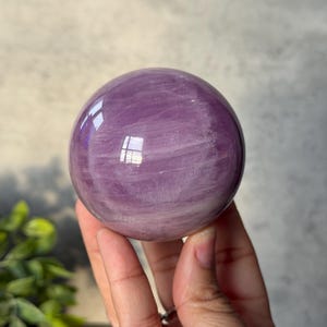 May include: A polished, spherical crystal ball featuring a translucent lavender and light purple hue. The surface displays natural internal veining and cloudy white inclusions throughout the stone, with a smooth, reflective finish that catches light.
