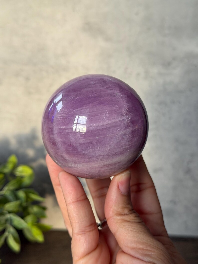 May include: A polished, spherical crystal ball featuring a translucent lavender and light purple hue. The surface displays natural internal veining and cloudy white inclusions throughout the stone, with a smooth, reflective finish that catches light.