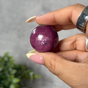 May include: A polished, deep magenta sphere, held in a hand. The gemstone has a smooth, reflective surface with subtle variations in color. The sphere is a rich, dark pink with hints of purple.
