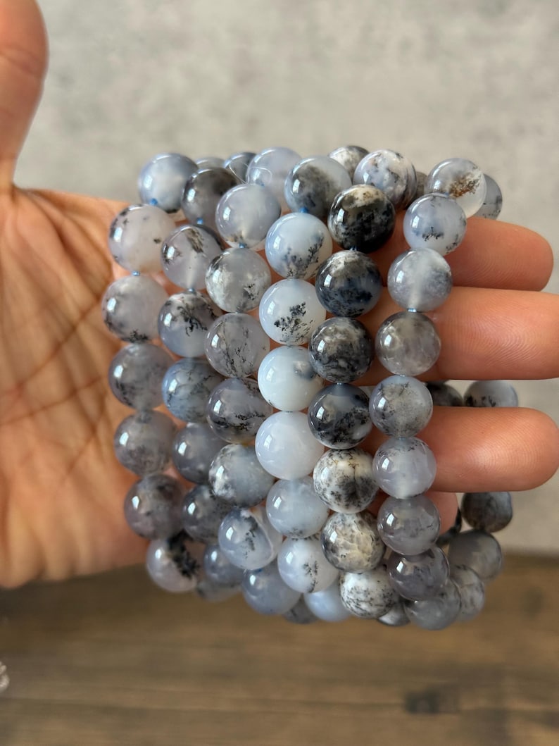 May include: A stack of gray and white dendritic agate bead bracelets. The beads are round and have a natural, organic look.