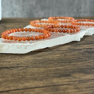 May include: Four orange sunstone beaded bracelets stacked on a white surface. The bracelets are made of small, round beads.