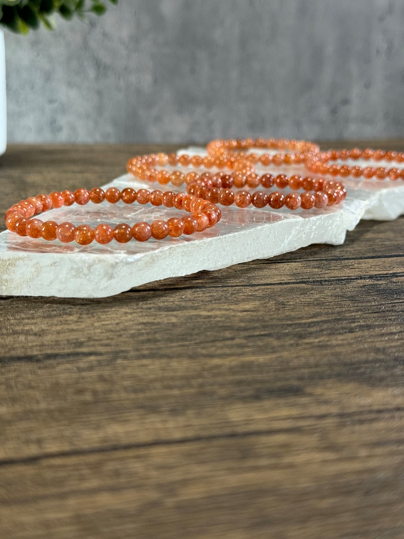 May include: Four orange sunstone beaded bracelets stacked on a white surface. The bracelets are made of small, round beads.