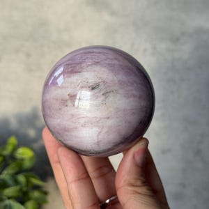 May include: A polished, spherical stone featuring a smooth surface with swirling patterns of light purple, lavender, and white, accented by subtle dark grey inclusions and a reflective sheen that highlights its rounded, orb-like form.