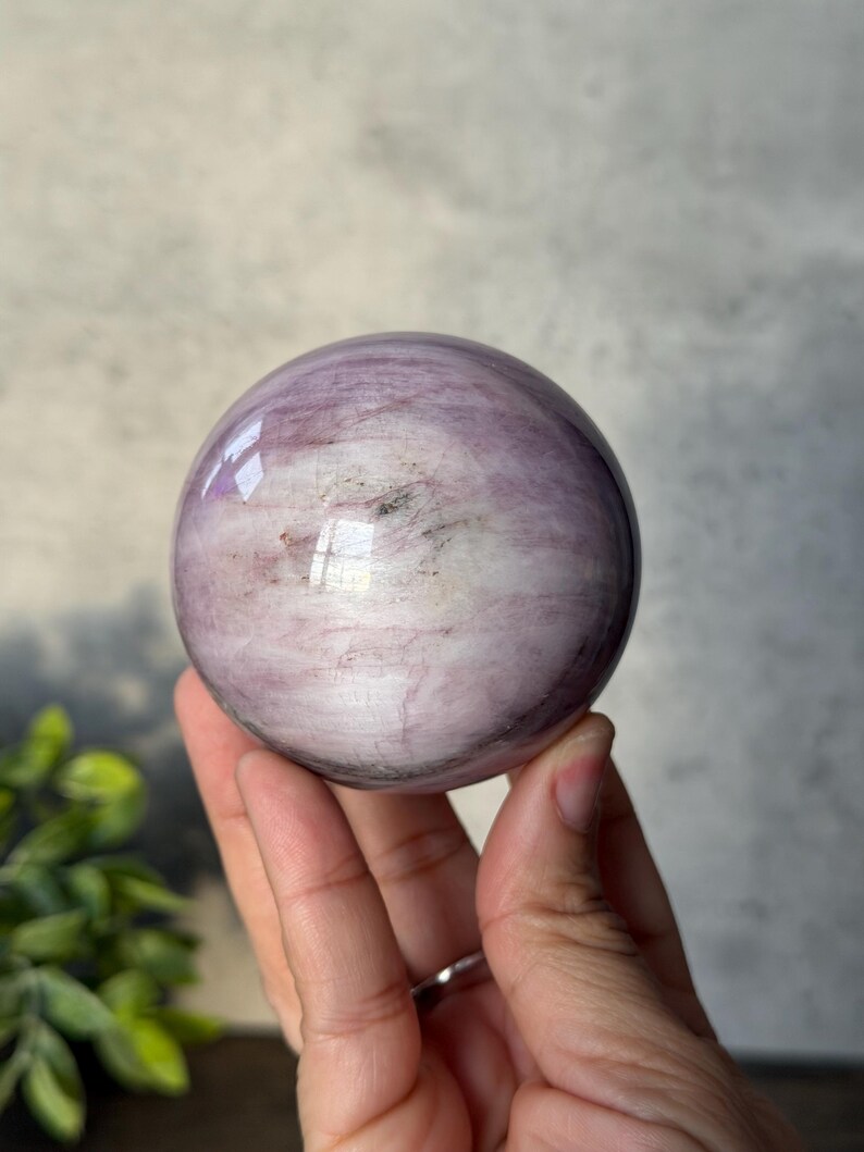 May include: A polished, spherical stone featuring a smooth surface with swirling patterns of light purple, lavender, and white, accented by subtle dark grey inclusions and a reflective sheen that highlights its rounded, orb-like form.
