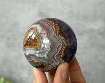 May include: A polished spherical stone featuring intricate concentric bands of white, gray, brown, and deep purple. The surface is smooth and reflective, showcasing natural mineral layers with varied textures and earthy color tones throughout.