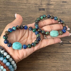 May include: Two beaded bracelets with a green and blue gemstone pattern. One bracelet has a larger, light green gemstone charm.