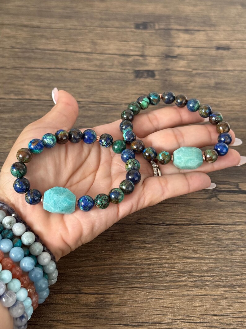 May include: Two beaded bracelets with a green and blue gemstone pattern. One bracelet has a larger, light green gemstone charm.