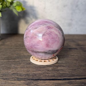 May include: A polished spherical stone featuring swirling patterns of light pink, magenta, and dark grey veins. The sphere rests on a small, circular, light-coloured wooden stand with a sunburst-like cutout design along its outer edge.