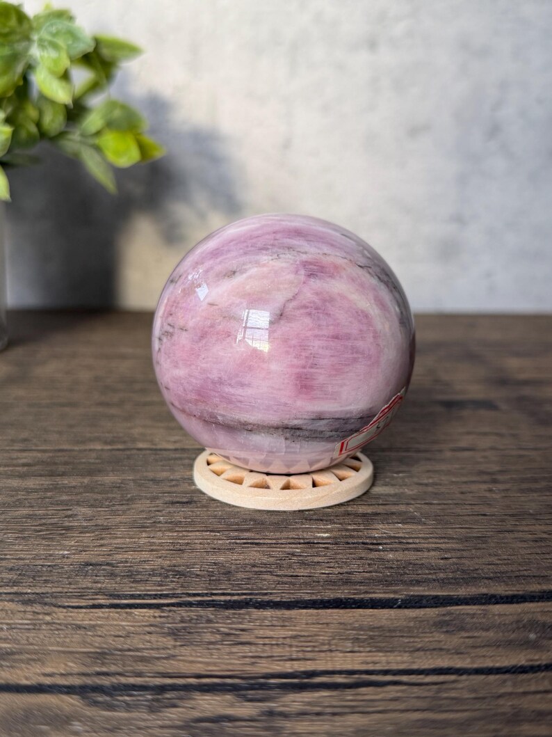 May include: A polished spherical stone featuring swirling patterns of light pink, magenta, and dark grey veins. The sphere rests on a small, circular, light-coloured wooden stand with a sunburst-like cutout design along its outer edge.