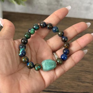 May include: A beaded bracelet with green, blue, and brown stones. The bracelet has a larger, light green stone in the center.